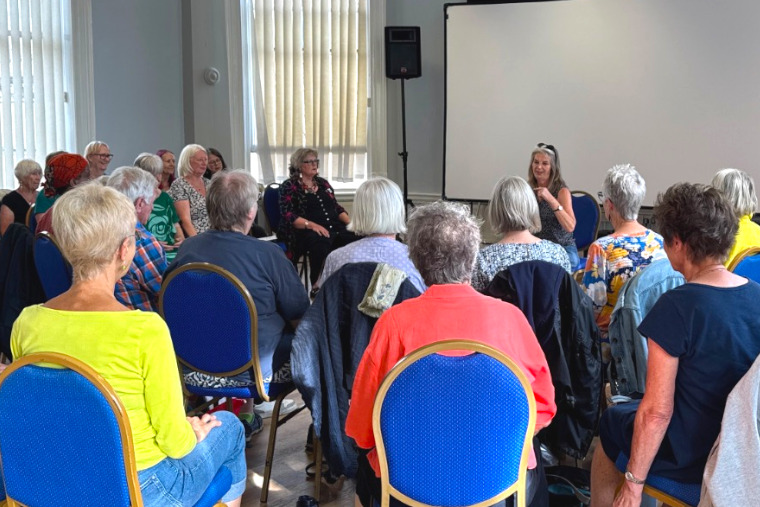 Website Chorus Rehearsal Image A group of men and women aged over 60 sitting in a rehearsal room with their backs to camera. Chorus Director, Em Whifield-Brooks, sits opposite them as she directs.